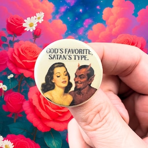 Puede incluir: Un botón redondo con el texto "GOD'S FAVORITE, SATAN'S TYPE." presenta una ilustración vintage de una mujer y un diablo. El botón se sostiene contra un fondo de rosas rosas y un cielo colorido.