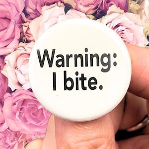 May include: A round, white button with the text "Warning: I bite" in black, held against a backdrop of pink and purple roses. The button is a simple, bold statement.