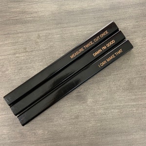 May include: Three black carpenter pencils with gold lettering. The pencils are stacked on top of each other. The text on the pencils reads: "Measure Twice, Cut Once", "Damn I'm Good", and "I Can Make That".