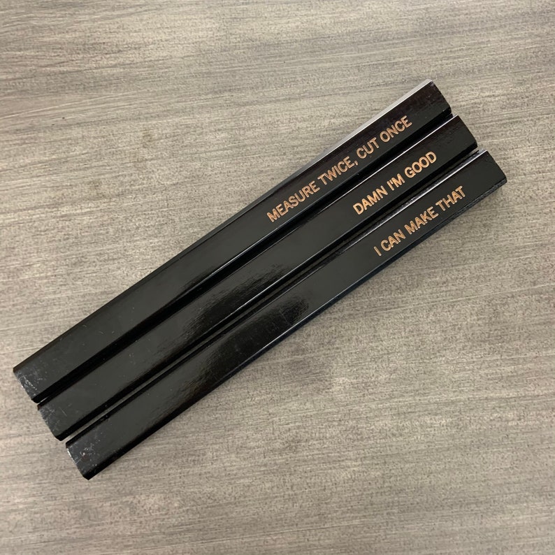 May include: Three black carpenter pencils with gold lettering. The pencils are stacked on top of each other. The text on the pencils reads: "Measure Twice, Cut Once", "Damn I'm Good", and "I Can Make That".