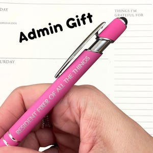 Puede incluir: Un bolígrafo rosa con una punta de estilete plateada y el texto "Resident Fixer of All the Things" grabado en el lateral. El bolígrafo se sostiene en una mano sobre un fondo blanco con las palabras "Admin Gift" escritas en negro encima del bolígrafo.