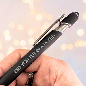 May include: Black pen with silver accents and a pocket clip. The pen has the phrase "DID YOU PUT IN A TICKET?" printed in white. The pen is held in a hand.