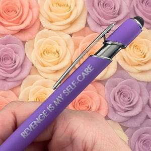 May include: A purple pen with the text "REVENGE IS MY SELF-CARE" in silver lettering. The pen has a silver clip and a black stylus tip. The background is a collection of pastel-colored roses.