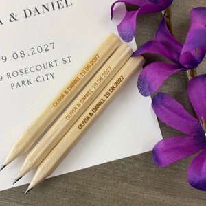 Op de afbeelding: Drie houten potloden met de tekst "Olivia & Daniel 19.08.2027" erop gegraveerd. De potloden liggen op een wit oppervlak met een paarse bloem op de achtergrond.