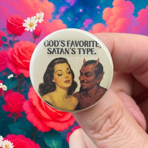 May include: A circular button displaying the text "GOD'S FAVORITE, SATAN'S TYPE." with a vintage illustration of a woman and a devil. The button is set against a backdrop of pink roses and a colourful sky.