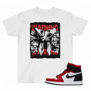 Puede incluir: Camiseta blanca con un diseño gráfico que presenta la palabra "WARNING" en rojo, junto con una ilustración en blanco y negro. Una zapatilla alta roja, blanca y negra también está en la imagen.