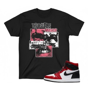 Puede incluir: Camiseta negra con la inscripción "TROUBLE MAKER" sobre un diseño gráfico rosa y blanco. Una zapatilla alta roja, blanca y negra se encuentra a la derecha de la camiseta, mostrando un diseño clásico.
