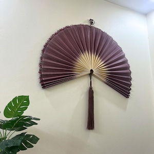 Puede incluir: Un gran abanico de pared semicircular con un diseño degradado que transita de un color morado ciruela intenso en los bordes exteriores a un tono crema claro cerca del punto de pivote central. Una borla larga a juego cuelga de la base.