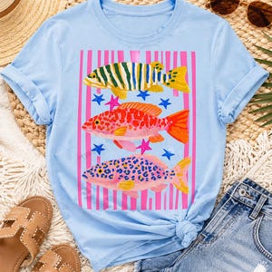Op de afbeelding: Lichtblauw T-shirt met een kleurrijk visontwerp. De afbeelding toont drie vissen in groen, oranje en roze, met roze strepen en blauwe sterren. Het shirt heeft opgerolde mouwen en een geknoopte zoom.