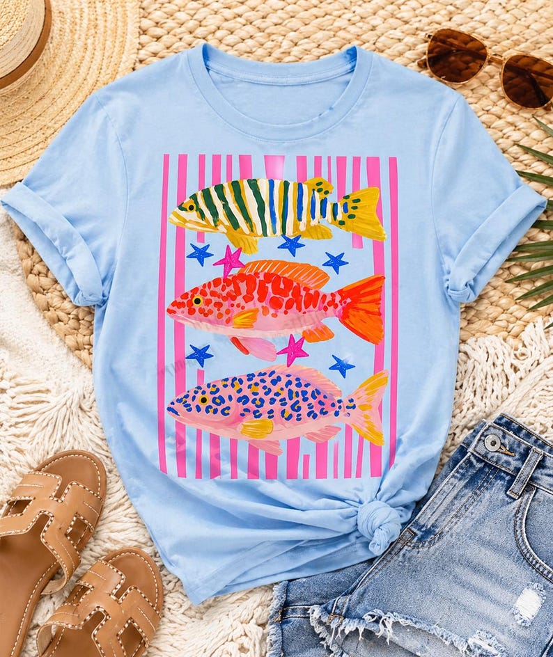 Op de afbeelding: Lichtblauw T-shirt met een kleurrijk visontwerp. De afbeelding toont drie vissen in groen, oranje en roze, met roze strepen en blauwe sterren. Het shirt heeft opgerolde mouwen en een geknoopte zoom.