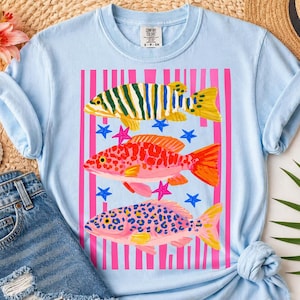 Op de afbeelding: Lichtblauw T-shirt met een kleurrijke afbeelding van drie vissen en sterren. De bovenste vis is groen en geel gestreept, de middelste vis is rood met oranje vinnen en de onderste vis is roze met blauwe stippen.