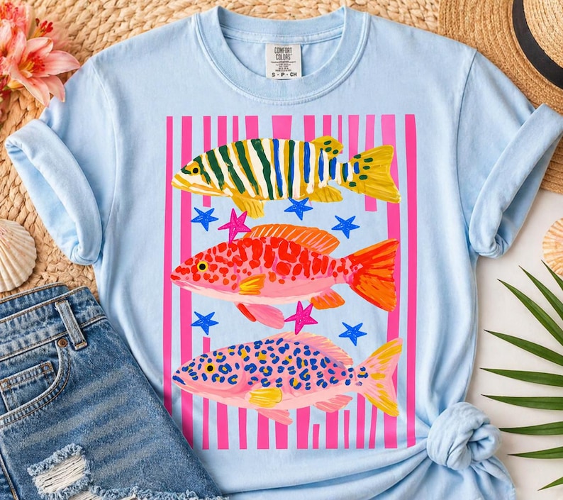 Op de afbeelding: Lichtblauw T-shirt met een kleurrijke afbeelding van drie vissen en sterren. De bovenste vis is groen en geel gestreept, de middelste vis is rood met oranje vinnen en de onderste vis is roze met blauwe stippen.