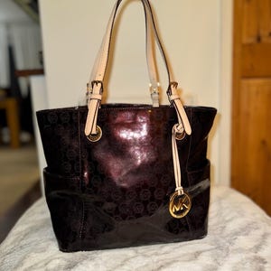 Op de afbeelding: Een glanzende, donker bordeauxrode tote bag met een herhalend patroon. De tas heeft twee zijvakken, crèmekleurige handvatten en goudkleurige hardware, waaronder een cirkelvormige logo-bedel. De tas staat op een zacht, lichtgekleurd oppervlak.