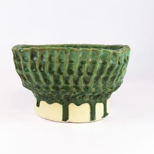 May include: A handcrafted ceramic bowl with a textured, green exterior and a cream-colored base. The bowl's surface has a unique, dimpled pattern, and the green glaze drips down the sides, creating a distinctive look. This decorative bowl is ideal for home decor.