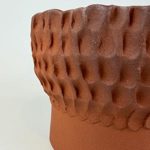 May include: A close-up of a handmade terracotta-colored ceramic pot. The pot has a textured, patterned surface with a series of rounded, raised shapes. The pot's rim is visible, and the background is a plain white.