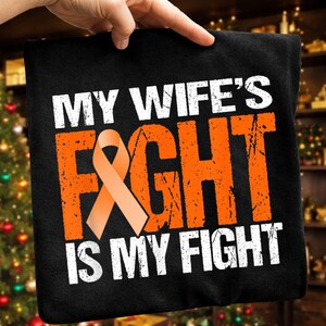 Puede incluir: Camiseta negra con el texto "MY WIFE'S FIGHT IS MY FIGHT" en letras blancas y naranjas. Una cinta naranja de concienciación está centrada en la camiseta. La camiseta está sujeta por una mano.