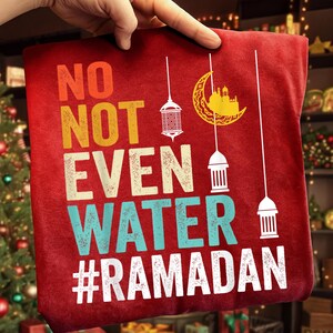 May include: Red t-shirt with the text "NO NOT EVEN WATER #RAMADAN" in orange, yellow, teal, and white. The design includes hanging lanterns and a crescent moon with a mosque silhouette.