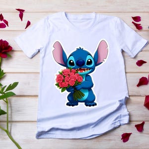 Puede incluir: Camiseta blanca con un personaje de dibujos animados azul sosteniendo un ramo de rosas rosas. El personaje tiene orejas grandes y una expresi&oacute;n amigable. La camiseta est&aacute; sobre una superficie de madera con p&eacute;talos de rosa esparcidos.