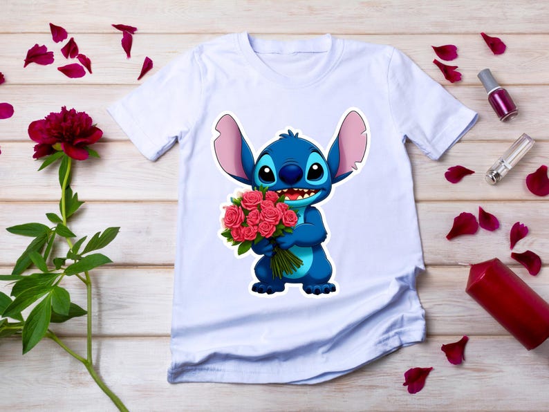 Puede incluir: Camiseta blanca con un personaje de dibujos animados azul sosteniendo un ramo de rosas rosas. El personaje tiene orejas grandes y una expresi&oacute;n amigable. La camiseta est&aacute; sobre una superficie de madera con p&eacute;talos de rosa esparcidos.