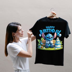 Puede incluir: Camiseta negra con un personaje de dibujos animados azul y la frase "HAPPY BIRTHDAY". El dise&ntilde;o incluye globos y una tarta de cumplea&ntilde;os. La camiseta la sostiene una persona.