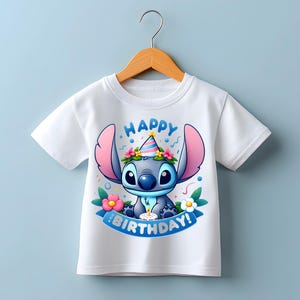 Puede incluir: Camiseta blanca con un personaje de dibujos animados con un gorro de fiesta y rodeado de flores. El texto "HAPPY BIRTHDAY!" es azul. La camiseta est&aacute; colgada en una percha de madera.