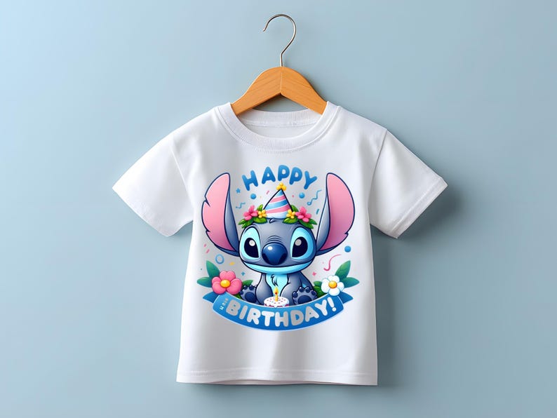 Puede incluir: Camiseta blanca con un personaje de dibujos animados con un gorro de fiesta y rodeado de flores. El texto "HAPPY BIRTHDAY!" es azul. La camiseta est&aacute; colgada en una percha de madera.
