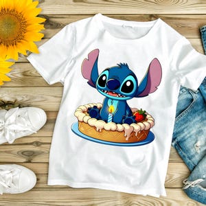 Puede incluir: Camiseta blanca con un personaje de dibujos animados, Stitch, sentado en una tarta con una vela encendida y una fresa. La imagen est&aacute; sobre una superficie de madera con girasoles, zapatillas blancas y vaqueros.
