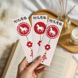 Può includere: Tre segnalibri decorativi con la silhouette di un cavallo bianco all'interno di un cerchio rosso, con caratteri cinesi. I segnalibri hanno uno sfondo a pois rosa e sono adornati con fiori rossi e un nodo decorativo.