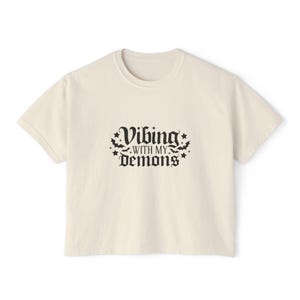 Op de afbeelding: Crèmekleurig T-shirt met de tekst "Vibing with my Demons" in zwarte gotische letters, versierd met vleermuis- en sterrenafbeeldingen. Het shirt heeft een ronde hals en korte mouwen, geschikt voor casual wear.