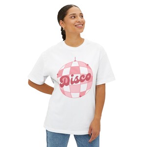 Può includere: T-shirt bianca con un motivo a palla da discoteca a scacchi rosa e bianchi e la parola "Disco" in corsivo. La maglietta è realizzata in un materiale morbido, adatta all'abbigliamento casual.