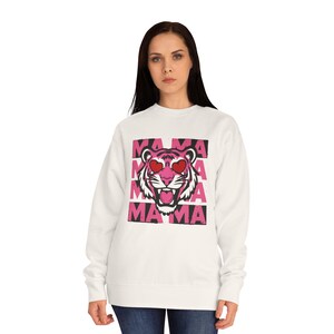 Può includere: Felpa bianca con un disegno di tigre rosa con occhi a forma di cuore e la parola "MAMA" in lettere rosa. Il disegno della tigre è centrato sulla parte anteriore del top a maniche lunghe e girocollo.