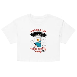Può includere: Top corto bianco con un design grafico raffigurante un UFO nero che fluttua sopra una donna con un vestito blu che legge un libro. Il testo recita "A BOOK A DAY keeps reality away BYE."