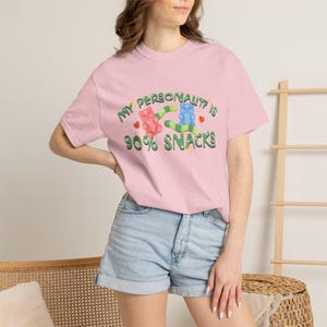 Può includere: T-shirt rosa chiaro con la scritta "MY PERSONALITY IS 90% SNACKS" in verde e giallo. La grafica include un orsetto gommoso rosa e blu e un serpente verde e blu. Abbinato a pantaloncini di jeans.