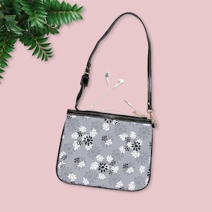 Può includere: Borsa a tracolla grigia con motivo floreale, cinturino e cerniera neri. La borsa presenta un motivo di fiori bianchi e neri. Gli auricolari bianchi sono visibili all'interno della borsa. La borsa è su sfondo rosa.