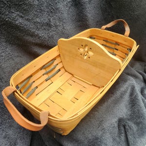 May include: A rectangular woven wood basket featuring a central divider with a decorative four-petal cutout design. The basket includes two tan leather handles attached to the ends and has a light natural wood finish with visible woven strips.