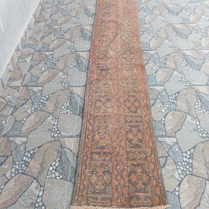 May include: A long, narrow rug with a detailed, repeating pattern in brown, orange, and green hues. The rug has a worn, vintage look and is laid on a stone tile floor. The rug's length is approximately 200 cm.
