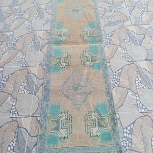 Puede incluir: Una alfombra pasillera rectangular con una base de color beige y patrones geométricos en tonos azul turquesa, azul claro y morado suave. La alfombra presenta un diseño simétrico con formas de diamante y motivos estilizados repetidos a lo largo.