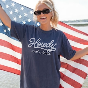 Navy blue t-shirt with the words "Howdy and Amen" in white script. The gal is holding an American flag with red and white stripes and white stars. The person is wearing sunglasses and gold hoop earrings.