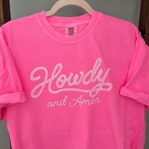 neon pink Comfort Colors t-shirt with the words "Howdy and Amen" in white script. The word "Howdy" is written in a rope-like font. The shirt is hanging on a white hanger.
