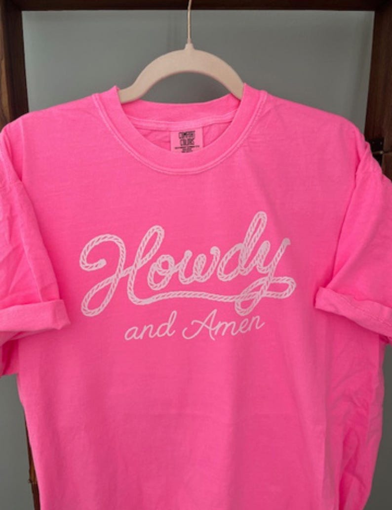 neon pink Comfort Colors t-shirt with the words "Howdy and Amen" in white script. The word "Howdy" is written in a rope-like font. The shirt is hanging on a white hanger.