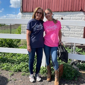 May include: A navy blue t-shirt and a bright pink t-shirt, both featuring the white cursive text Howdy and Amen printed on the front. The navy shirt has short sleeves and a crew neckline, while the pink shirt also has short sleeves and a crew neckline.