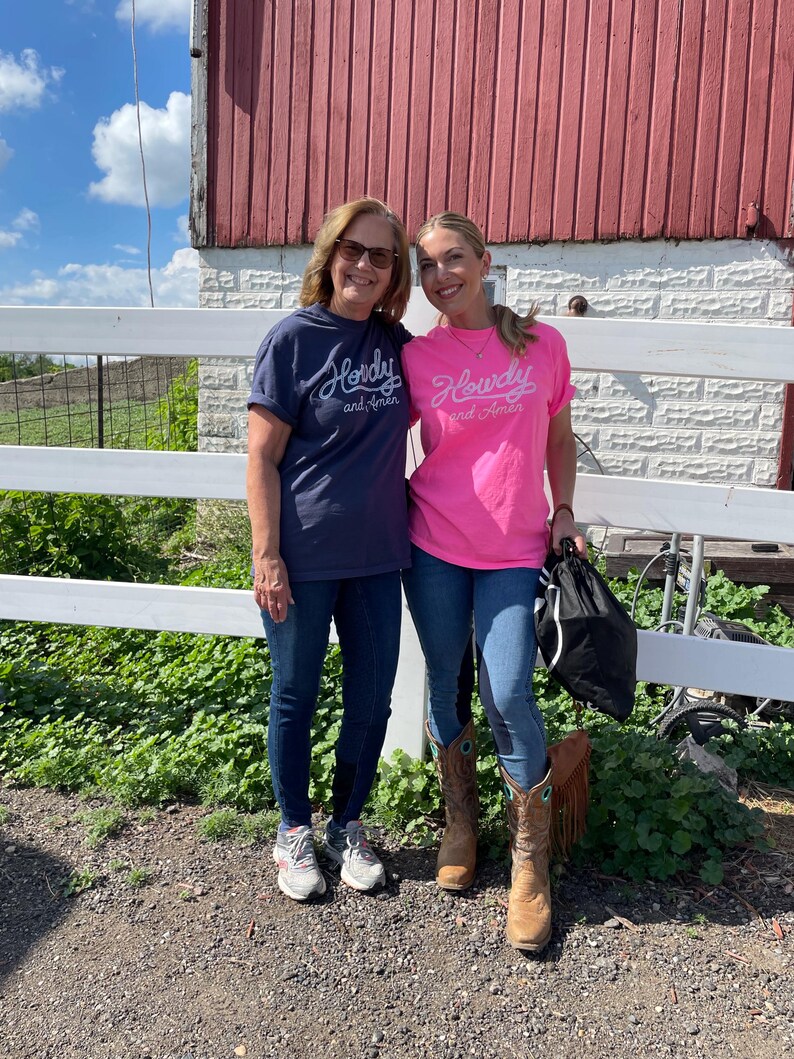 May include: A navy blue t-shirt and a bright pink t-shirt, both featuring the white cursive text Howdy and Amen printed on the front. The navy shirt has short sleeves and a crew neckline, while the pink shirt also has short sleeves and a crew neckline.