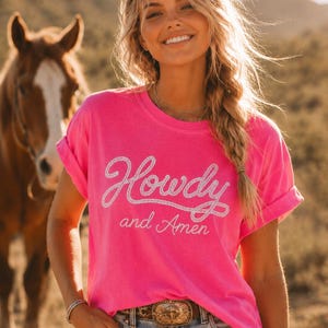 May include: A bright neon pink short-sleeved t-shirt featuring the white text Howdy and Amen printed on the front. The word Howdy is written in a stylized rope-like font, while the words and Amen appear in a thinner, cursive script below it.
