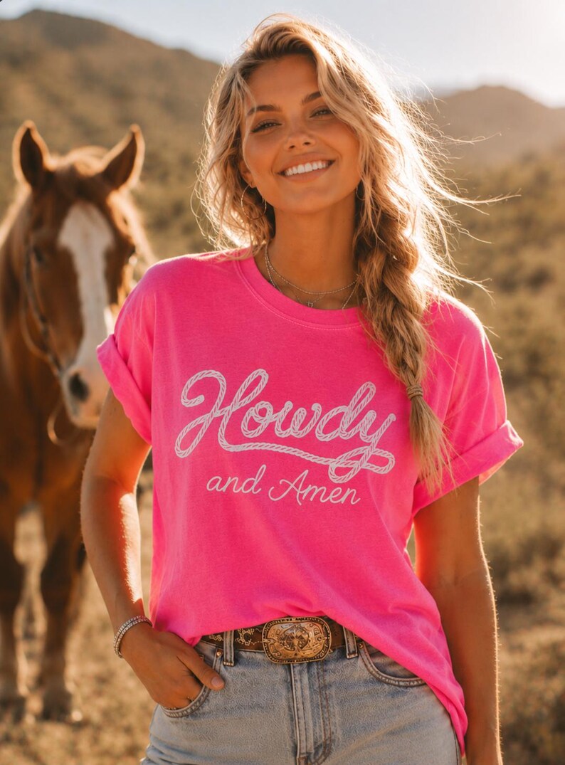 May include: A bright neon pink short-sleeved t-shirt featuring the white text Howdy and Amen printed on the front. The word Howdy is written in a stylized rope-like font, while the words and Amen appear in a thinner, cursive script below it.