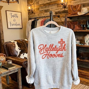 Może przedstawiać: Szara bluza z napisem "Hallelujah Hooves" w czerwonym skrypcie, z motywem krzyża. Bluza jest prezentowana na wieszaku w butiku. Z tyłu widnieje szyld z napisem "Faith & Freedom".