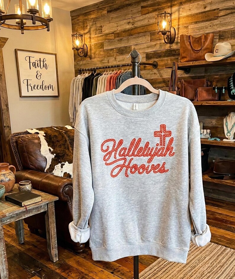 Może przedstawiać: Szara bluza z napisem "Hallelujah Hooves" w czerwonym skrypcie, z motywem krzyża. Bluza jest prezentowana na wieszaku w butiku. Z tyłu widnieje szyld z napisem "Faith & Freedom".