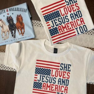 May include: White t-shirts with patriotic designs. One shirt features the text "SHE LOVES JESUS AND AMERICA TOO" with an American flag design. Another shirt has the same text. A light blue shirt has the text "WHITE & HALLELUJAH" with horse graphics.