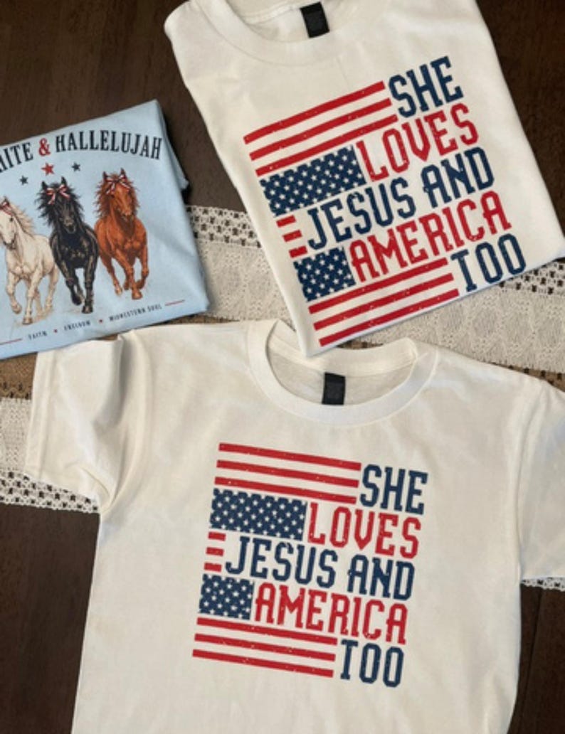 May include: White t-shirts with patriotic designs. One shirt features the text "SHE LOVES JESUS AND AMERICA TOO" with an American flag design. Another shirt has the same text. A light blue shirt has the text "WHITE & HALLELUJAH" with horse graphics.