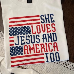 May include: White t-shirt with a distressed American flag design in red and blue. The text reads "SHE LOVES JESUS AND AMERICA TOO" in a bold, patriotic font. The shirt is made of a soft material.