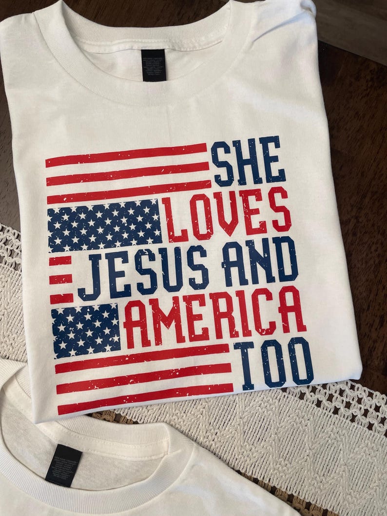 May include: White t-shirt with a distressed American flag design in red and blue. The text reads "SHE LOVES JESUS AND AMERICA TOO" in a bold, patriotic font. The shirt is made of a soft material.
